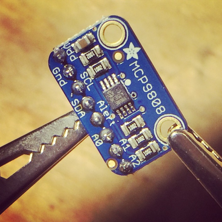 The MCP9808 sensor, a small blue Adafruit breakout board, held next to a house key for scale.